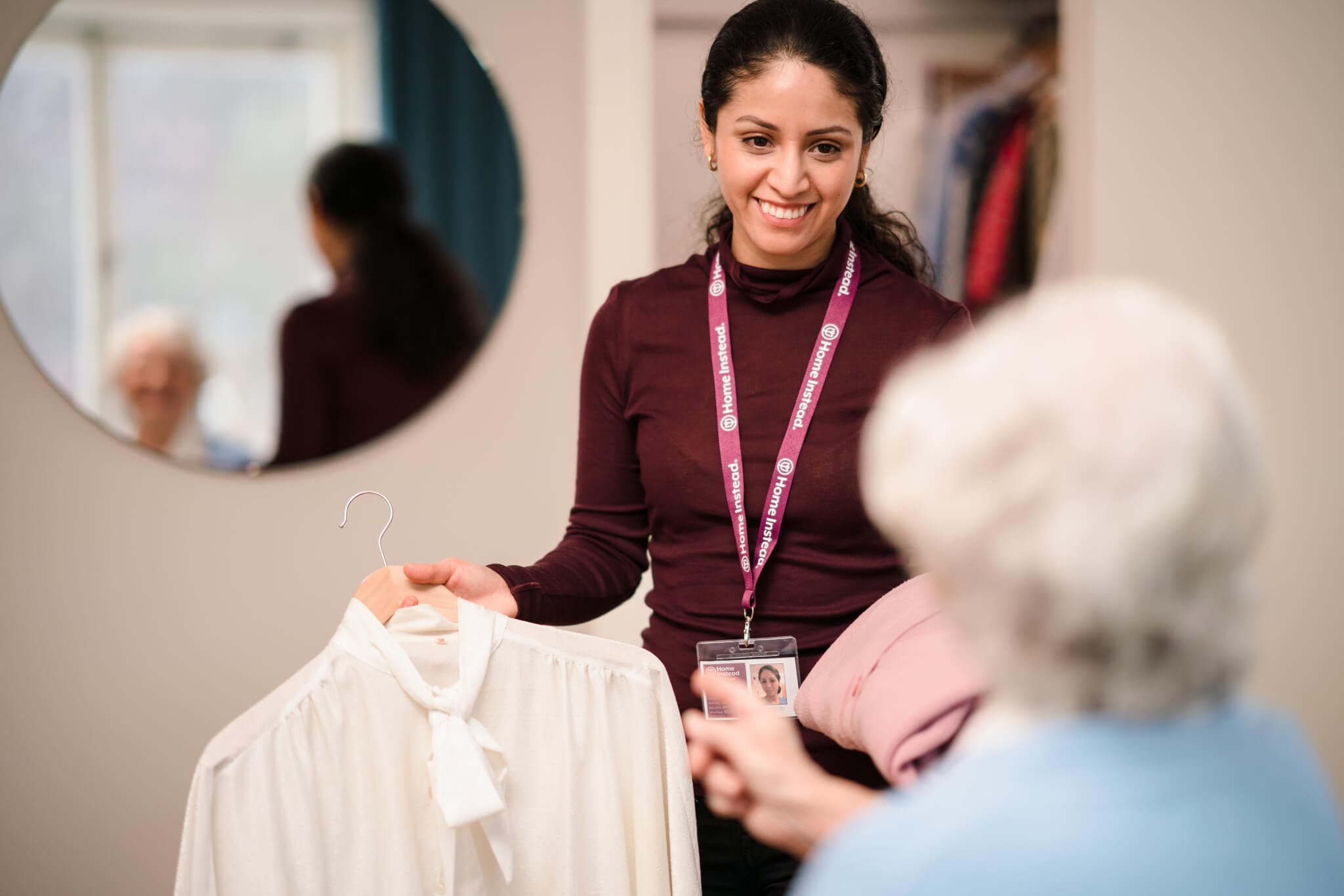 A caregiver smiles and shows a white blouse on a hanger to an elderly woman in a home setting. - Home Instead