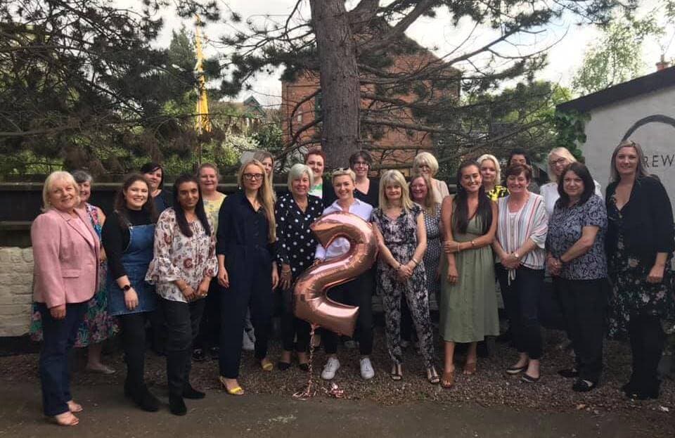 Group of people standing outside, one person holding a balloon shaped like the number 2, all smiling at the camera. - Home Instead