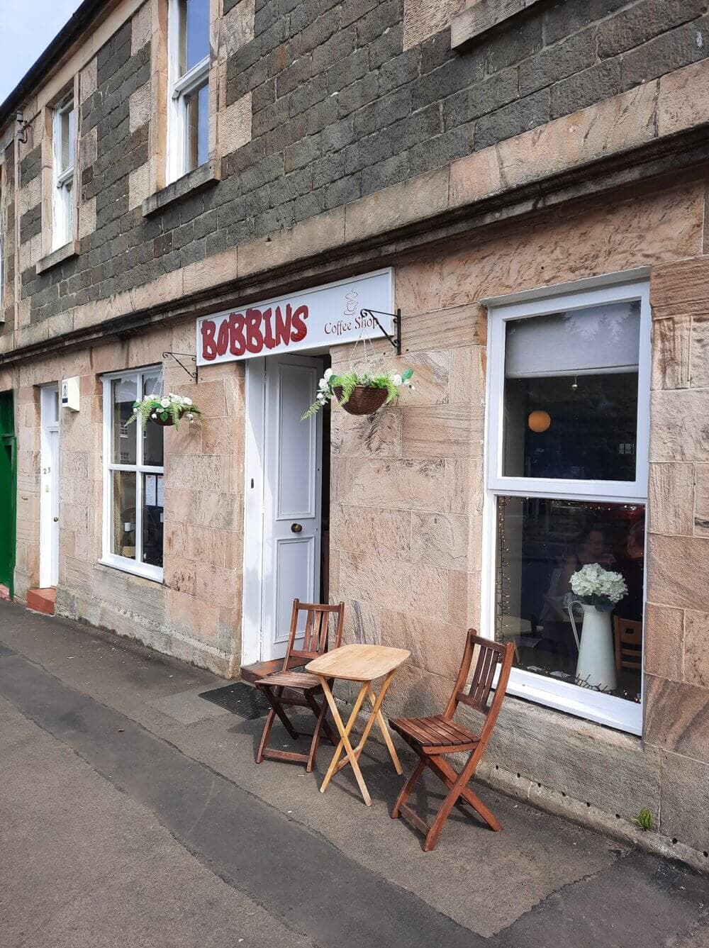 A small café with a sign that reads "Bobbins", two wooden chairs, and a table outside on a stone-paved sidewalk. - Home Instead Renfrewshire and Barrhead care at home services