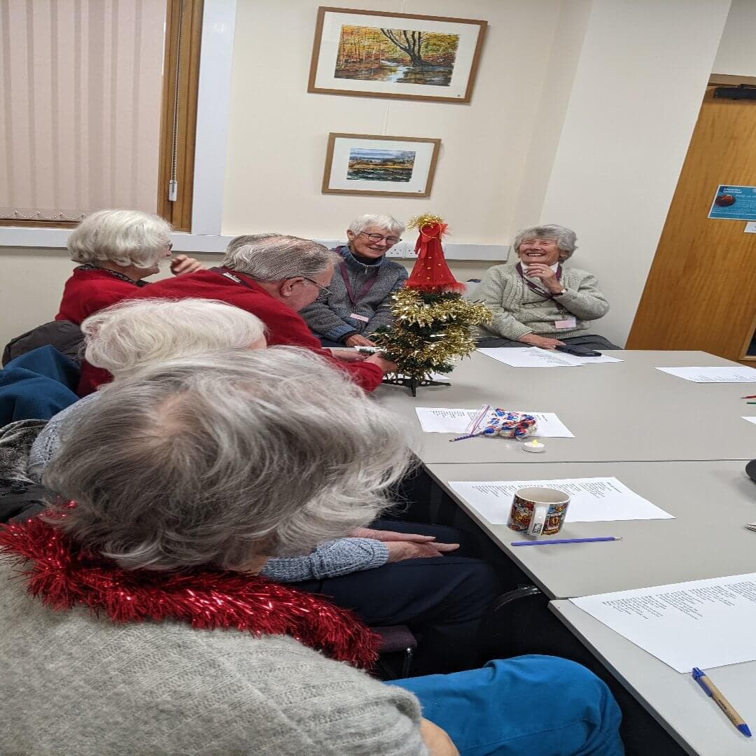 Group of older adults sitting around a table with papers, a small Christmas tree, and festive decorations in a meeting room. - Home Instead