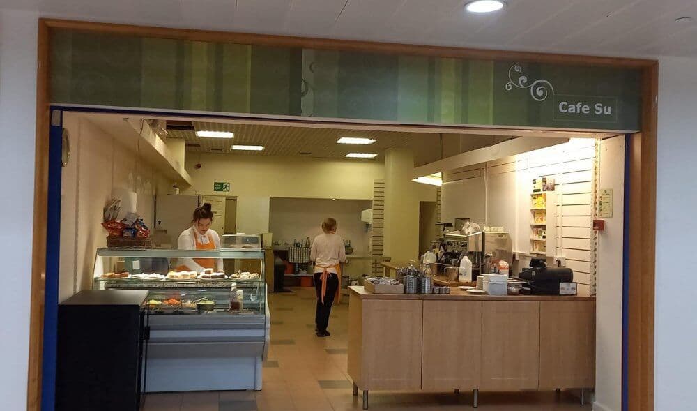 Two people working in a cafe with a counter displaying pastries and a sign that reads "Cafe Su" above the entrance. - Home Instead Renfrewshire and Barrhead care at home services
