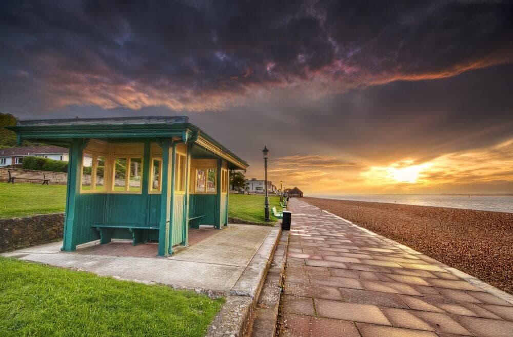 A seaside shelter along a paved path at sunset, with dramatic clouds and a grassy area to the left. - Home Instead