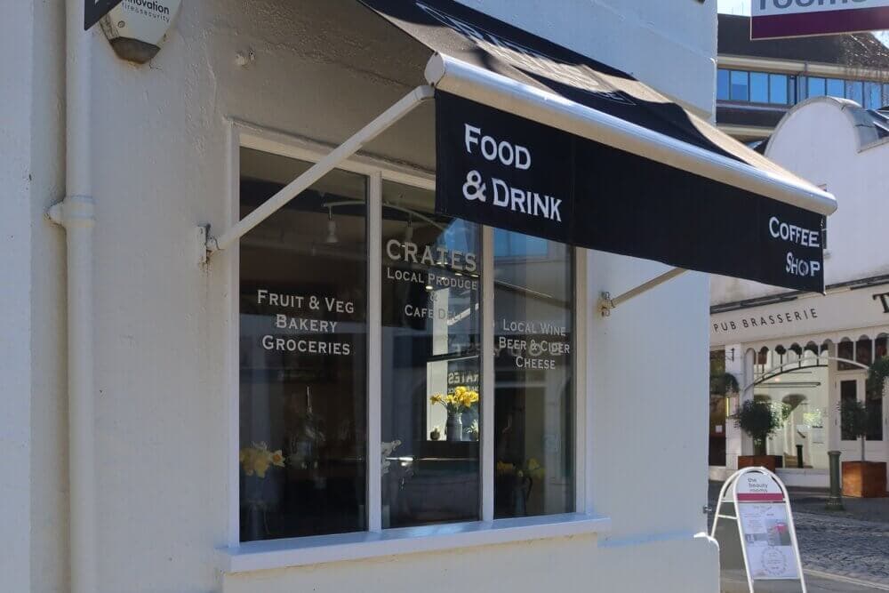 A shop with a black awning labeled "Food & Drink" and "Coffee Shop," with various items listed on the windows. - Home Instead