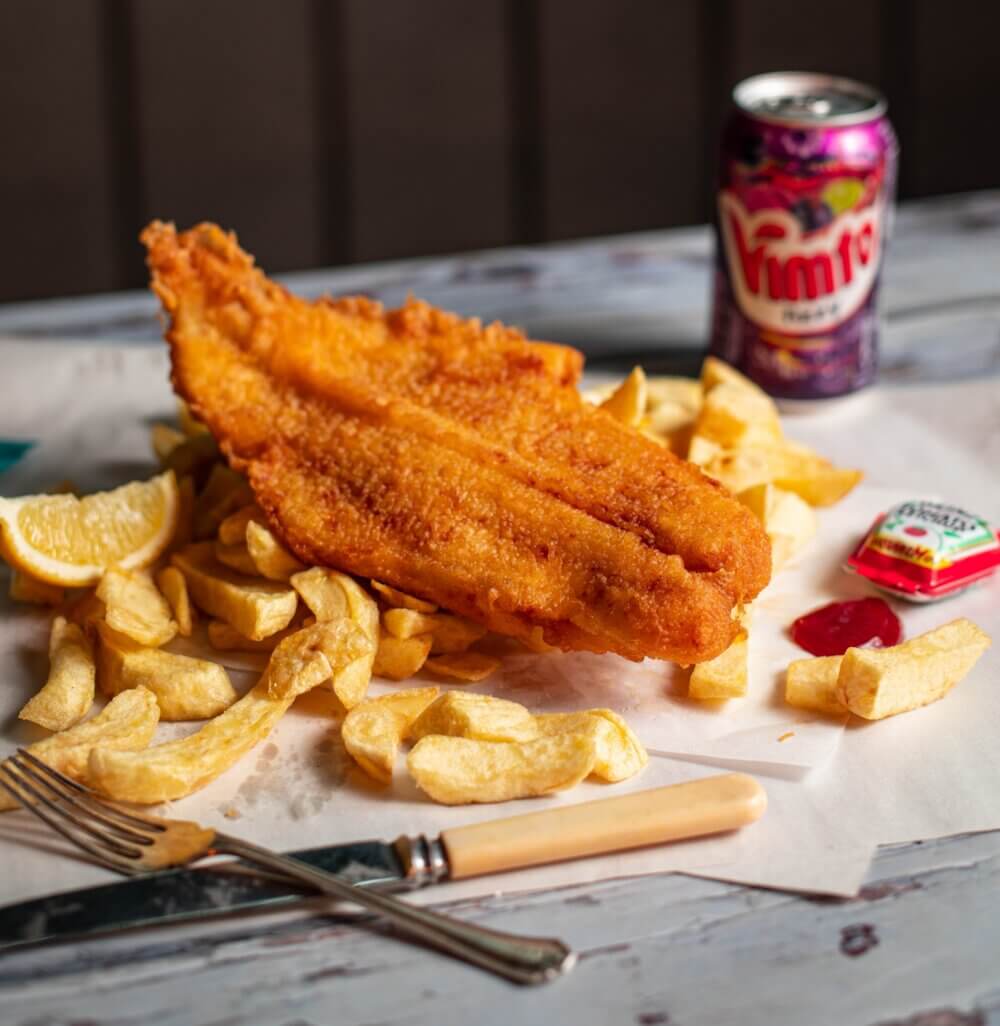 Enzo's - A plate of fish and chips with a lemon wedge, a fork, a knife, ketchup, and a can of Vimto soft drink. - Home Instead Renfrewshire and Barrhead care at home