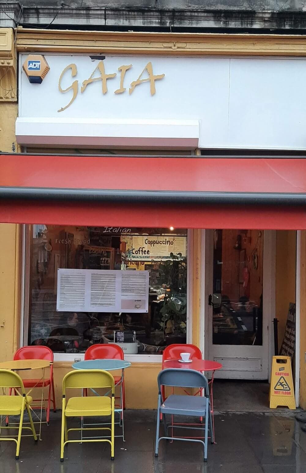 A small cafe with a red awning, colorful chairs, and tables on the sidewalk, with the sign "GAIA" above the entrance. - Home Instead