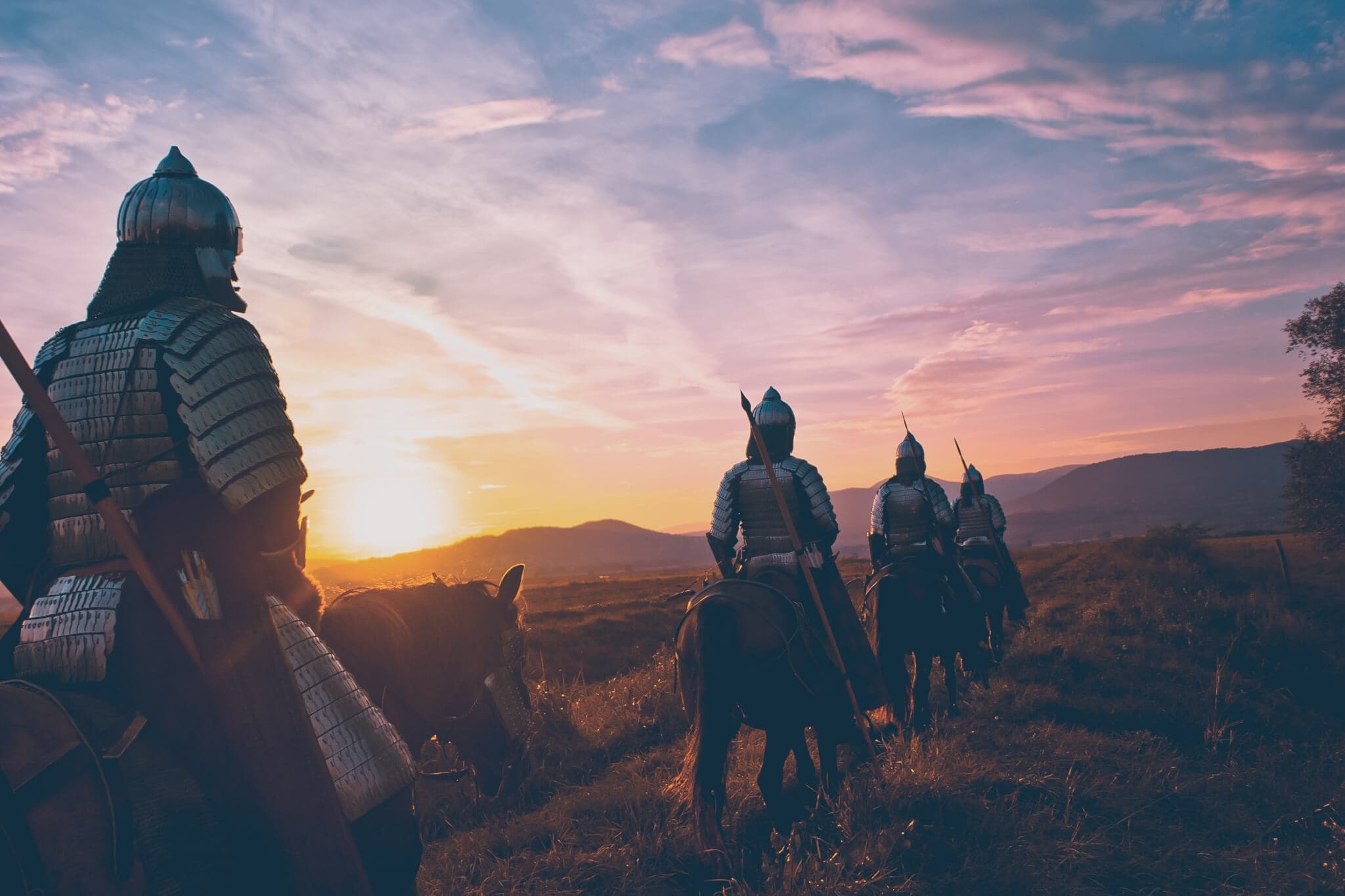 Four medieval knights on horseback ride towards a sunset over a grassy landscape with mountains in the distance. - Home Instead