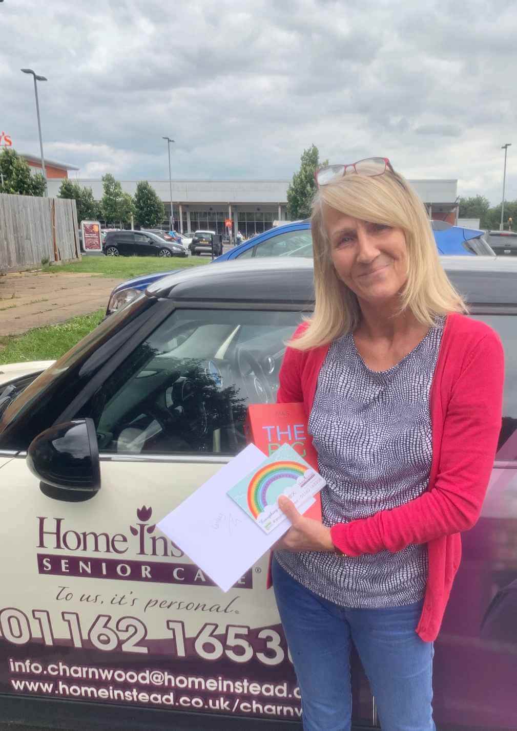 A woman holds a thank-you card and book, standing by a car with "Home Instead Senior Care" branding. - Home Instead