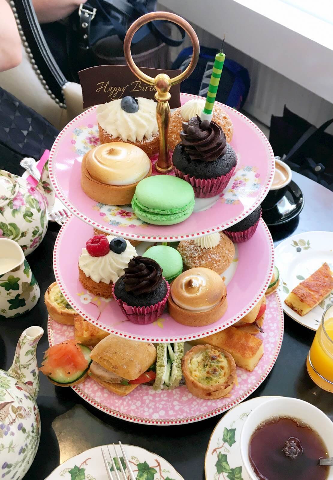 A three-tiered stand filled with assorted pastries, cupcakes, macarons, sandwiches, and desserts, with tea cups around. - Home Instead