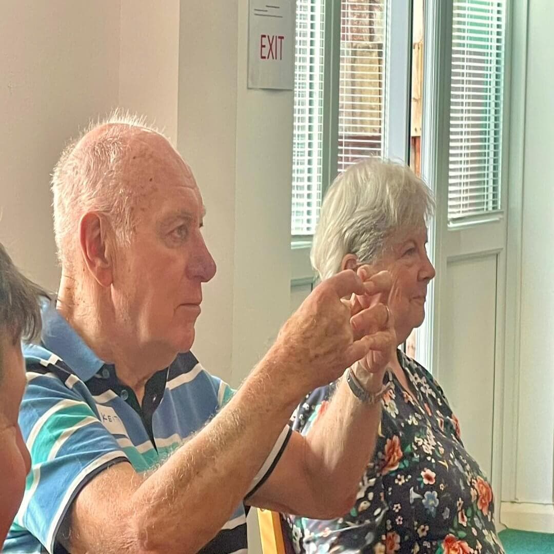An elderly man and woman sitting indoors, attentively listening. The man holds his hands up. An exit sign is visible. - Home Instead