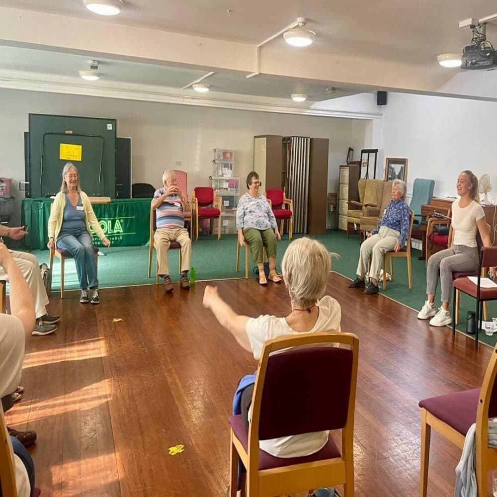 A group of elderly individuals participating in a seated exercise session in a bright, spacious room. - Home Instead