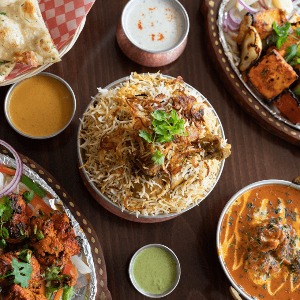 Overhead view of various Indian dishes, including biryani, naan, kebabs, and curry, arranged on a wooden table. - Home Instead