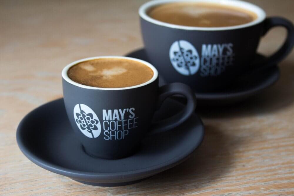 Two black coffee cups with "May's Coffee Shop" logo, one small, one large, placed on saucers on a wooden table. - Home Instead Renfrewshire and Barrhead home care services