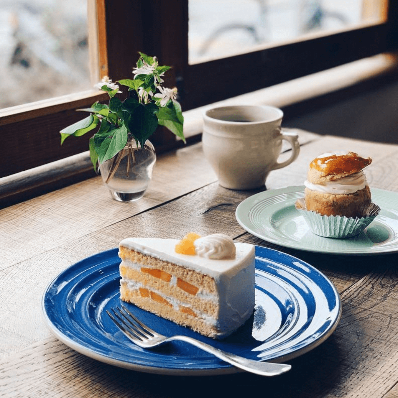 Slice of fruit layer cake on a blue plate, cream pastry on a green plate, a cup, and a vase on a wooden table by a window. - Home Instead