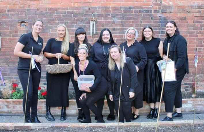 A group of women dressed in black stands in front of a brick wall, smiling and holding various containers. - Home Instead