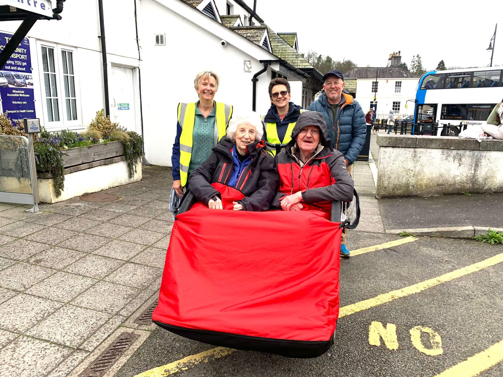 Two elderly people sit in a red rickshaw, smiling, with three people standing behind them, outside a building. - Home Instead