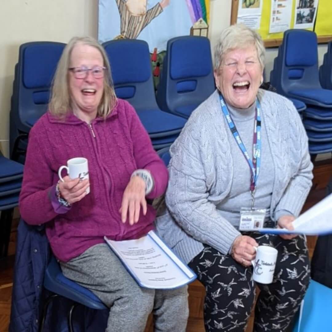 Two elderly women sit on chairs, holding mugs, and laughing together in a room with stacked chairs in the background. - Home Instead