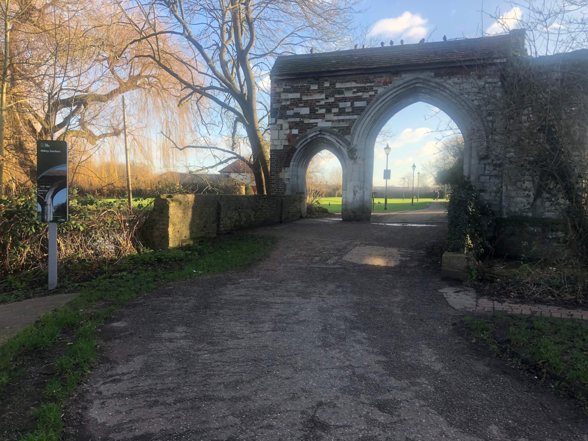 An old stone archway with a path leading to a sunny park, with bare trees and a signpost on the left side. - Home Instead