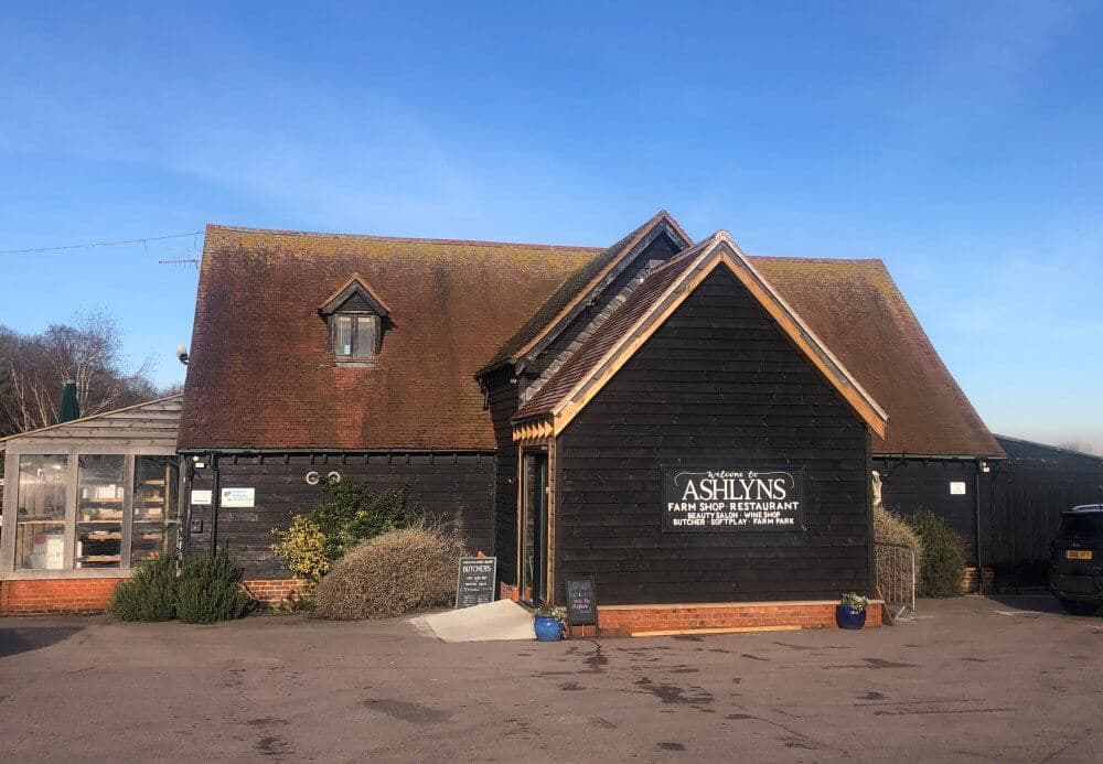 A rustic building with a sloped roof houses Ashlyns Farm Shop and Restaurant, fronted by a parked car and plants. - Home Instead