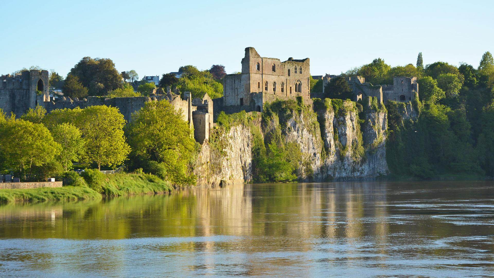 Medieval castle ruins situated on a cliff overlooking a calm river, surrounded by lush green trees under a clear sky. - Home Instead