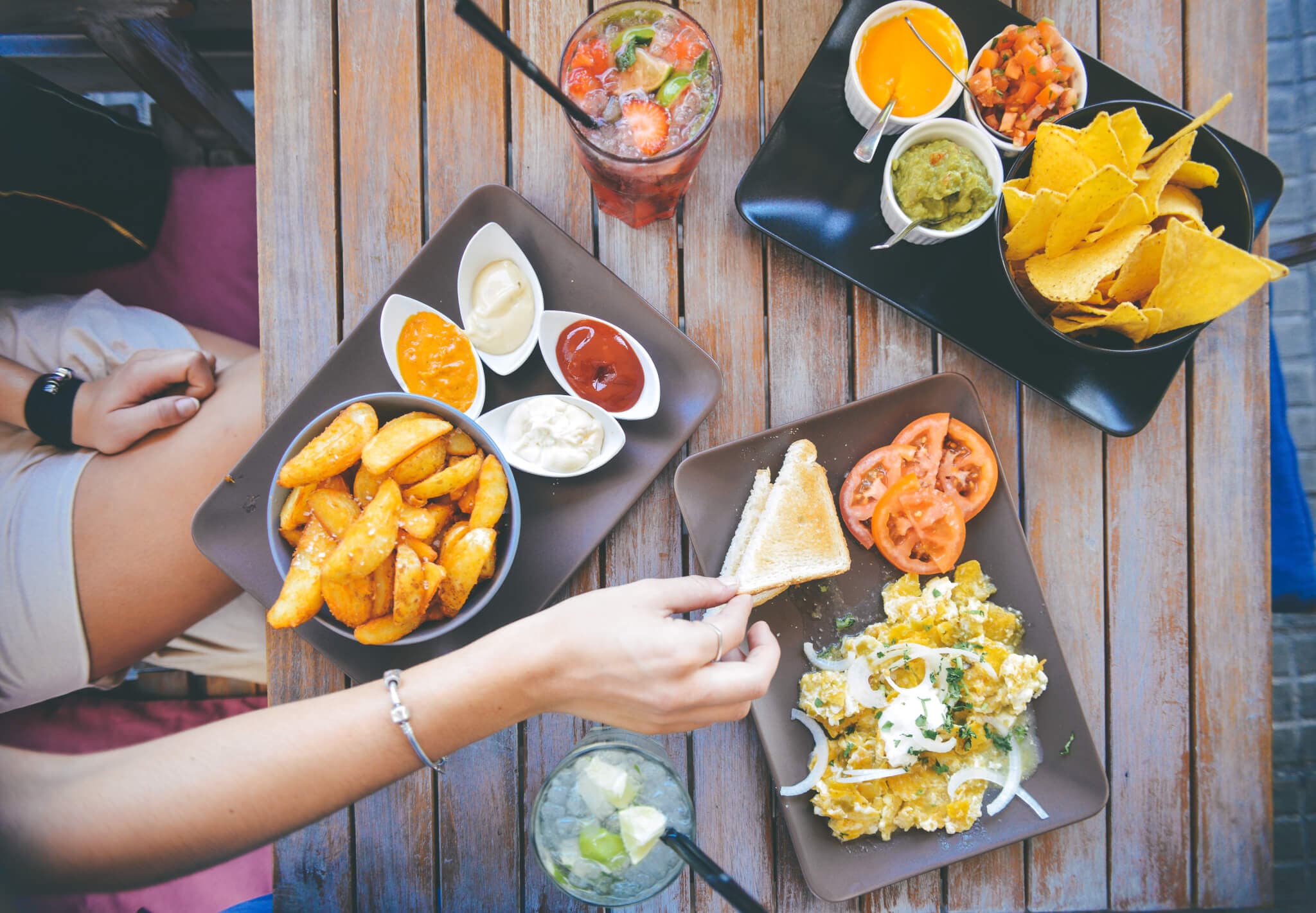Overhead view of a table with nachos, dipping sauces, potato wedges, salad, drinks, and someone reaching for food. - Home Instead