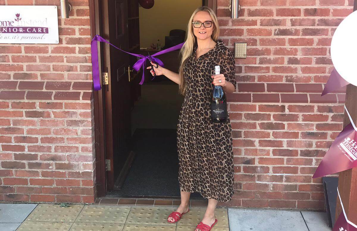 A woman in a leopard-print dress cuts a purple ribbon at an event, holding a bottle. She stands in front of a brick building. - Home Instead