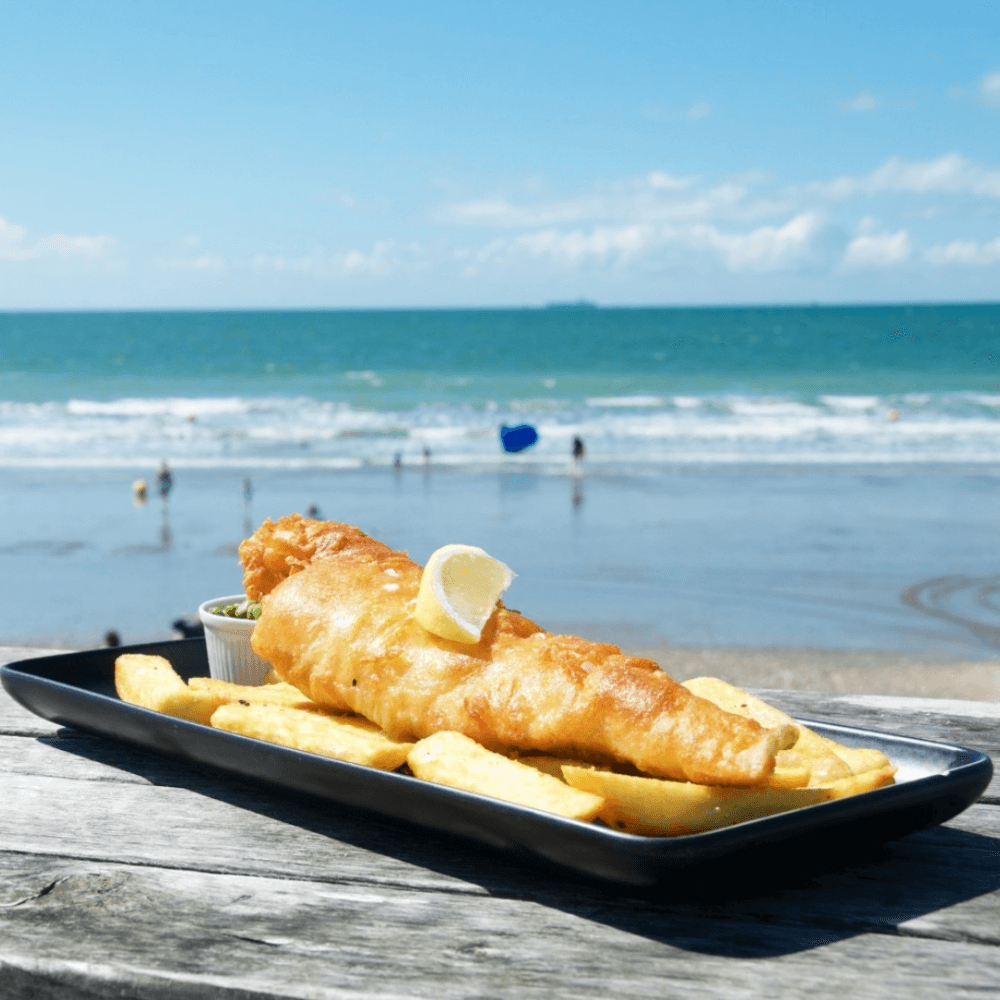 Fish and chips with a lemon wedge on a black plate, placed on a wooden table by the beach with waves and people in the background. - Home Instead