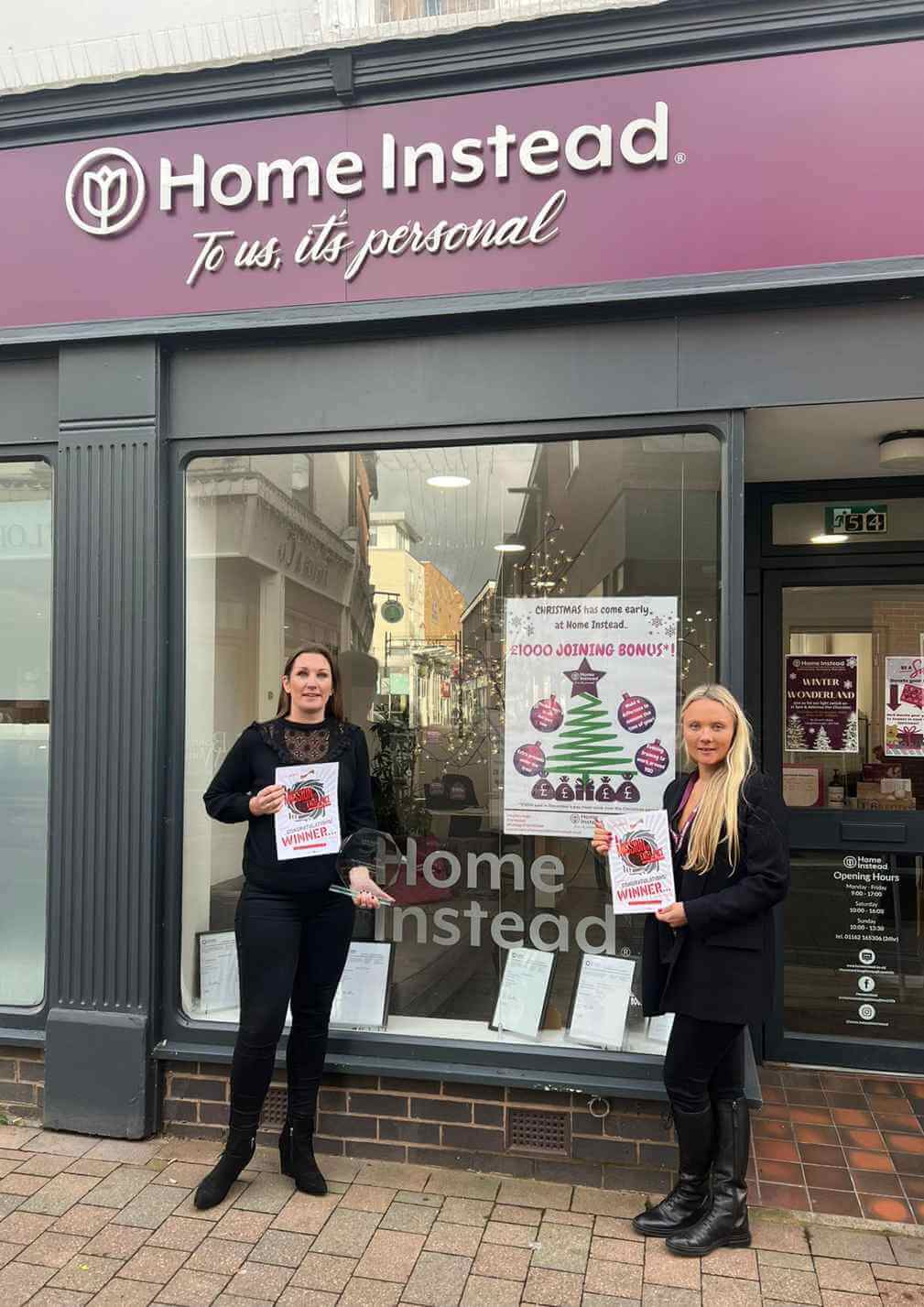 Two people standing in front of a Home Instead storefront, holding flyers. The store window displays posters and signs. - Home Instead