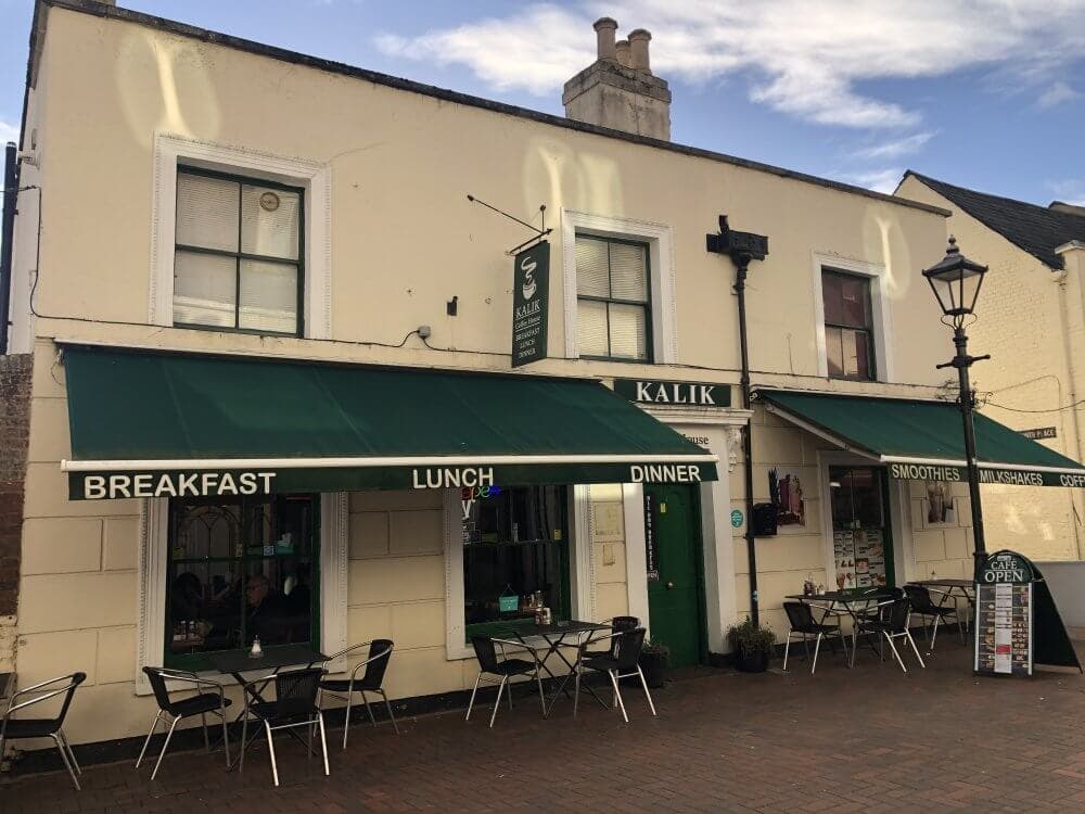 Street view of a café with green awnings, outdoor seating, and signs advertising breakfast, lunch, dinner, smoothies, and milkshakes. - Home Instead