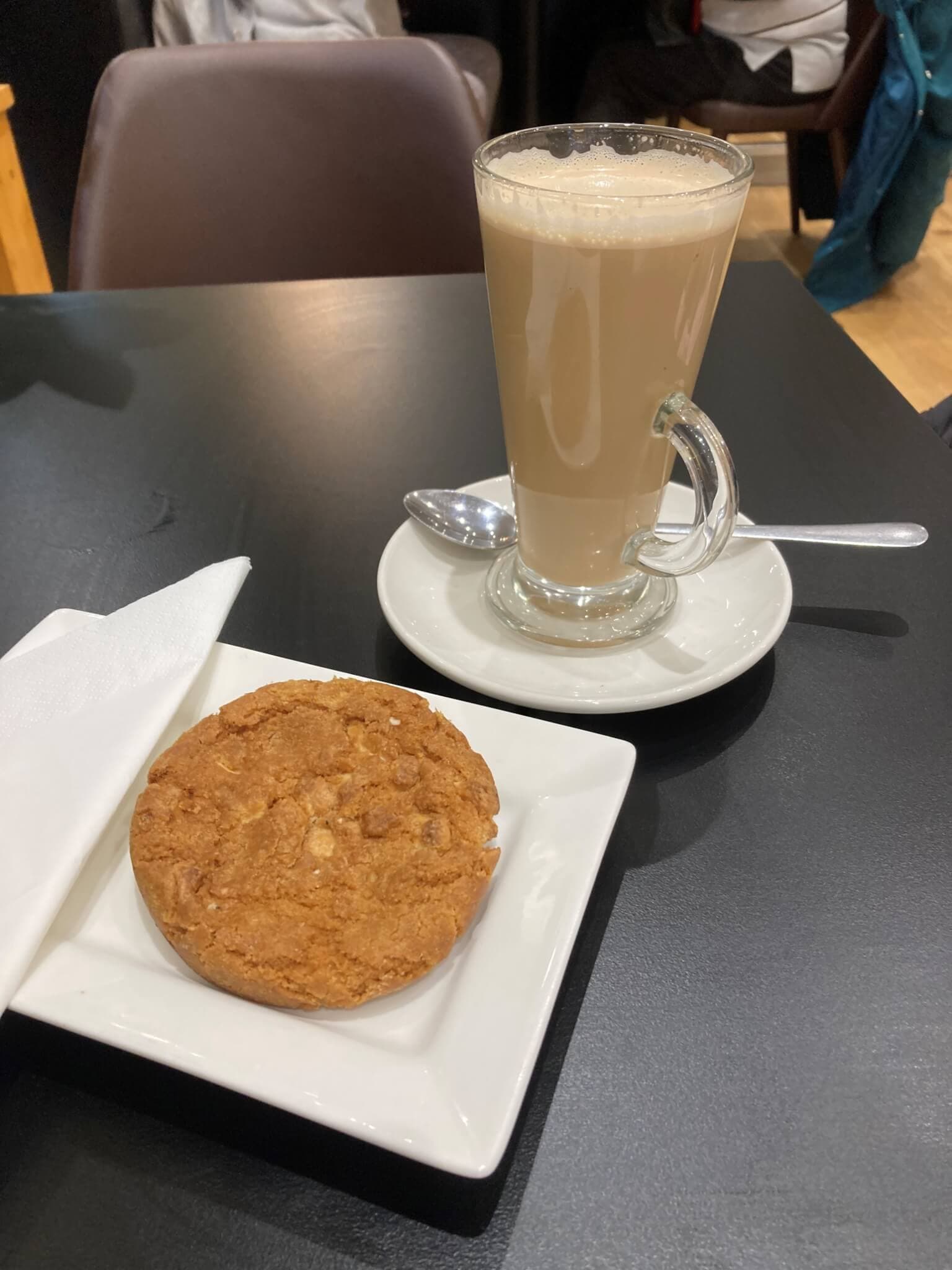 A latte in a glass mug with a spoon on a saucer next to a cookie on a plate with a napkin on a black table. - Home Instead