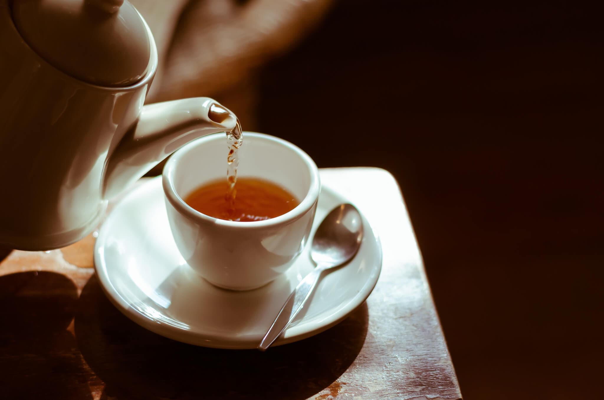 A teapot pours tea into a white cup on a saucer with a spoon, placed on a wooden table in warm, soft lighting. - Home Instead