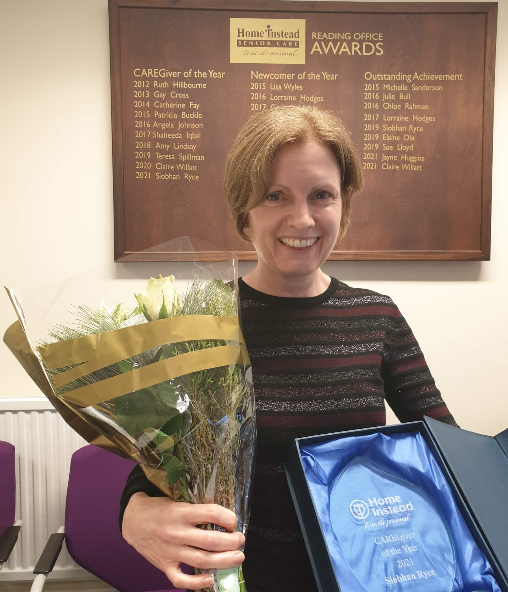 A smiling woman holds a bouquet and an award. A list of past award winners is displayed on the wall behind her. - Home Instead