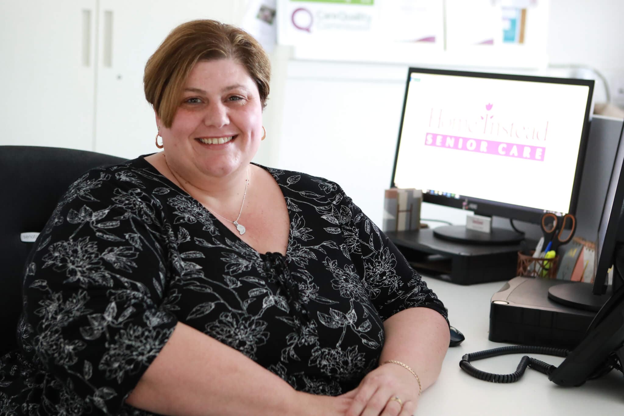 A woman wearing a floral blouse sits smiling at a desk with a computer displaying "Home Instead Senior Care. - Home Instead