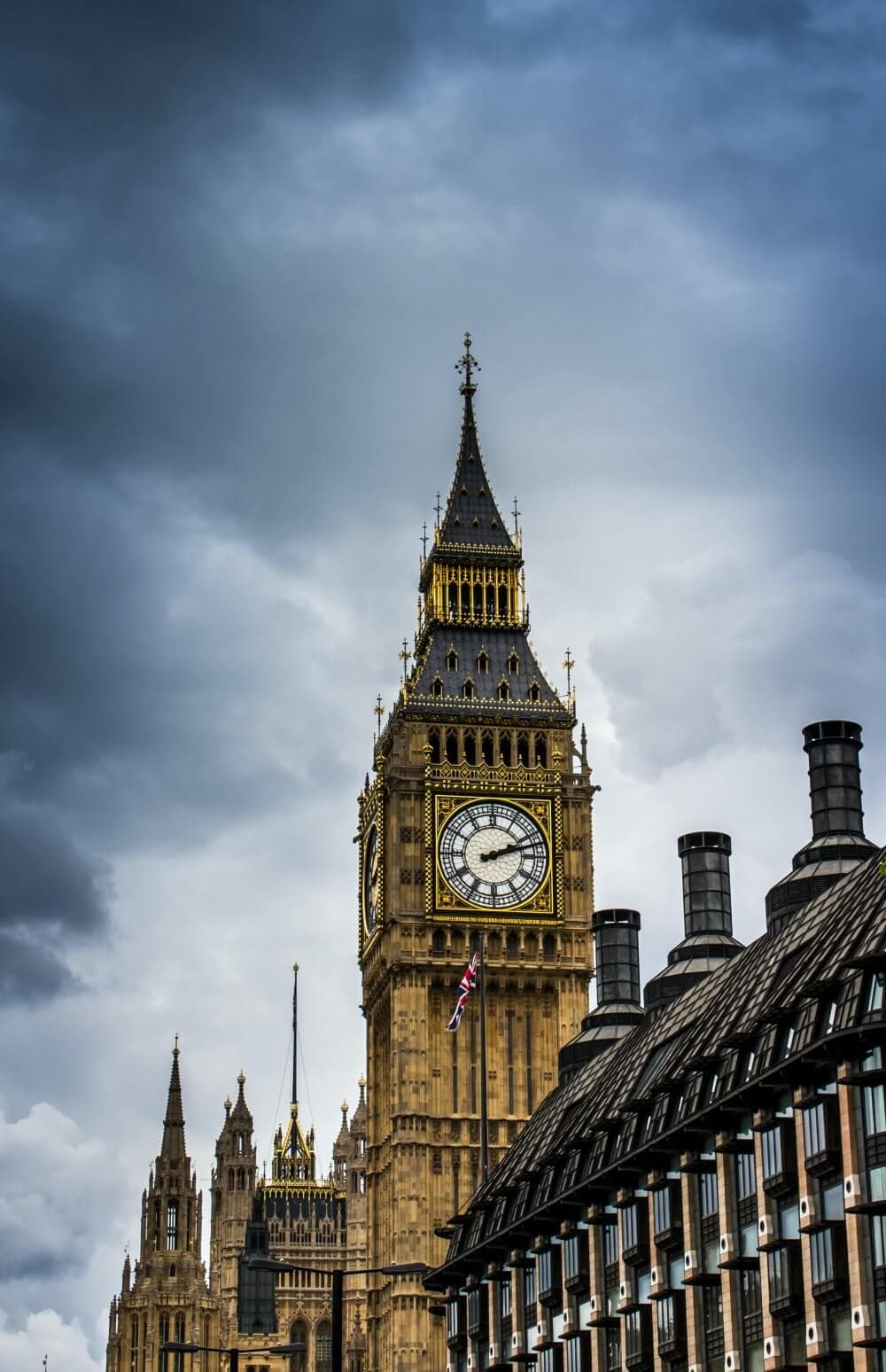 The Elizabeth Tower with Big Ben clock in London against a cloudy sky, with part of a building in the foreground. - Home Instead