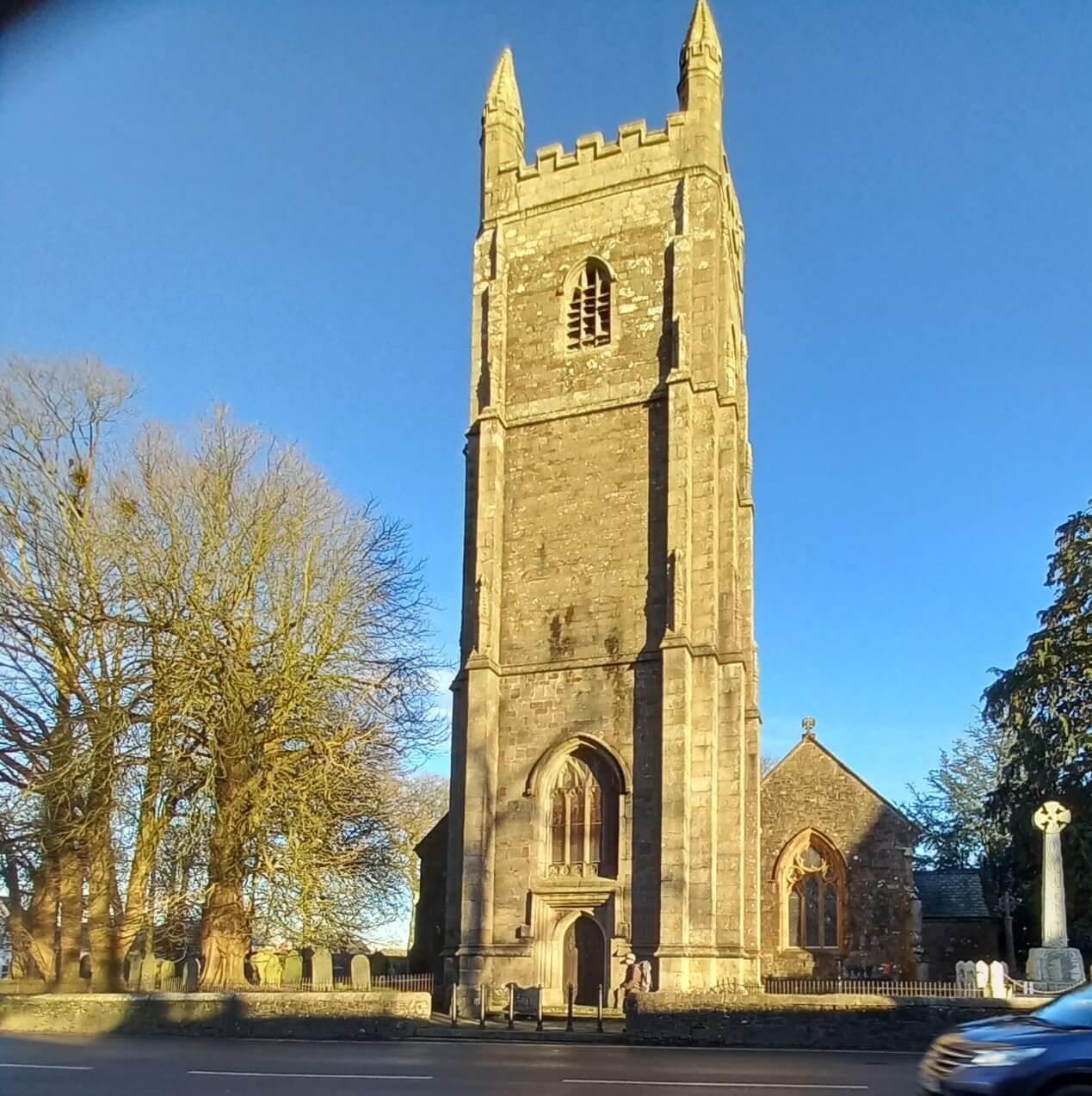 A stone church with a tall tower and clock, surrounded by trees, on a clear day with a partly cloudy blue sky. - Home Instead