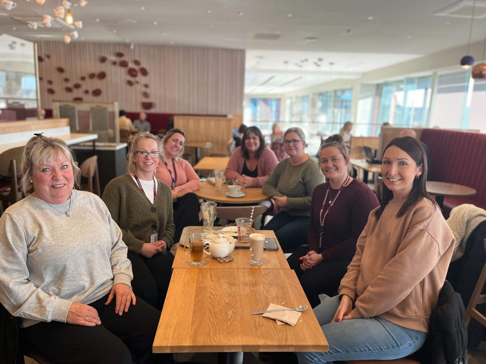 A group of seven women smiling and sitting around a table in a cozy café. - Home Instead