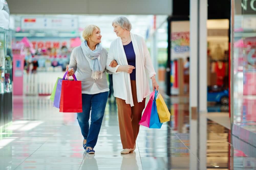 Two elderly women walking in a mall, holding hands and carrying colorful shopping bags, smiling at each other. - Home Instead