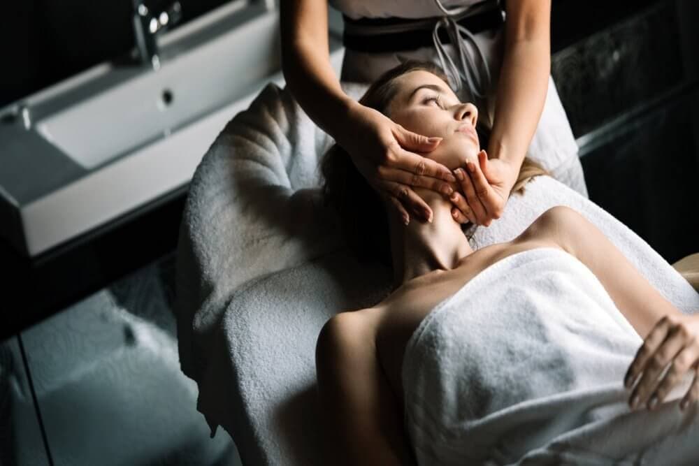 Person relaxing on a spa bed with a towel wrapped around, receiving a face massage in a dimly lit room. - Home Instead Bournemouth & Christchurch