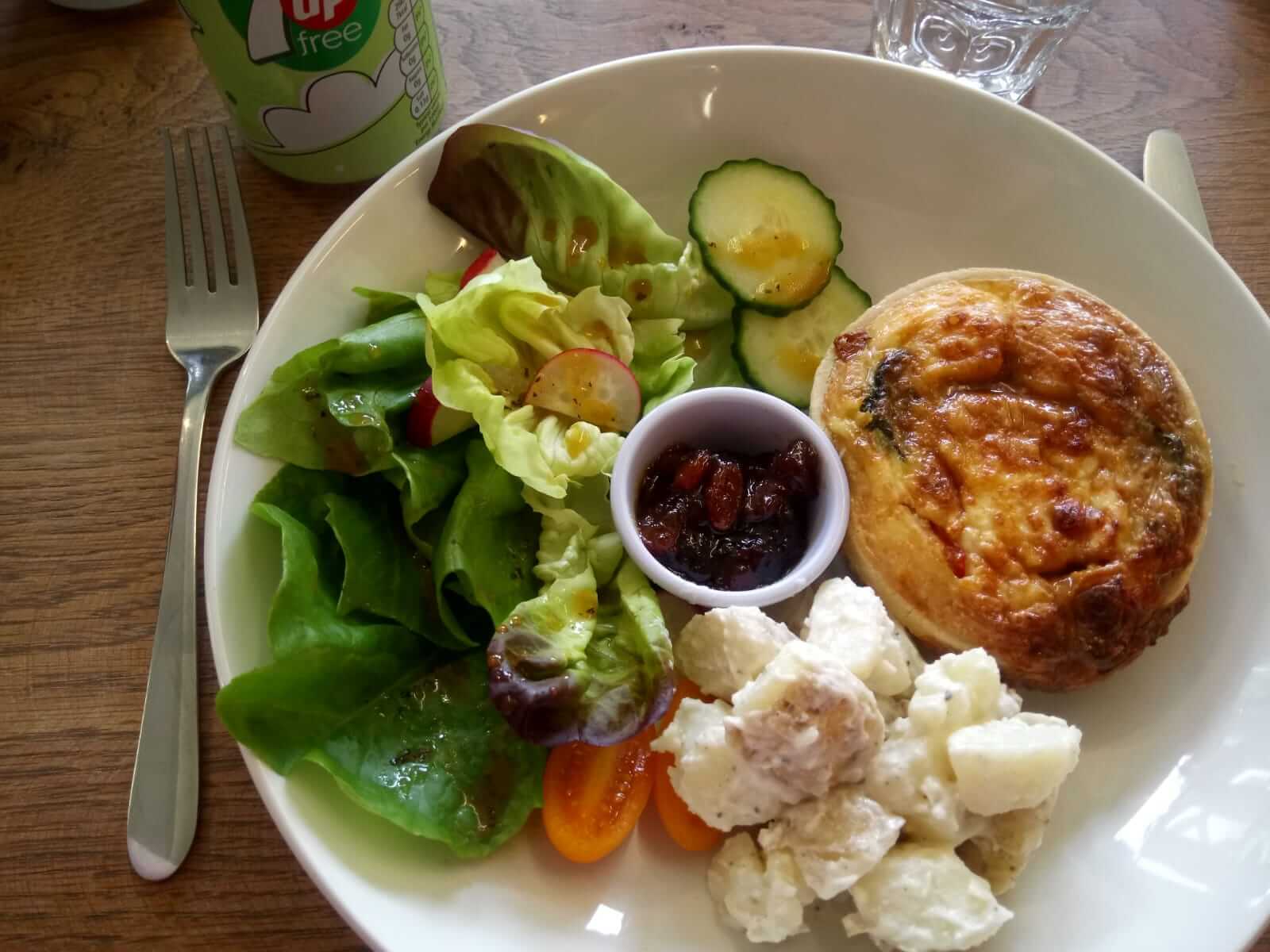 A plate with salad, potato salad, a small pie, and a side of chutney, accompanied by a drink in a green cup. - Home Instead