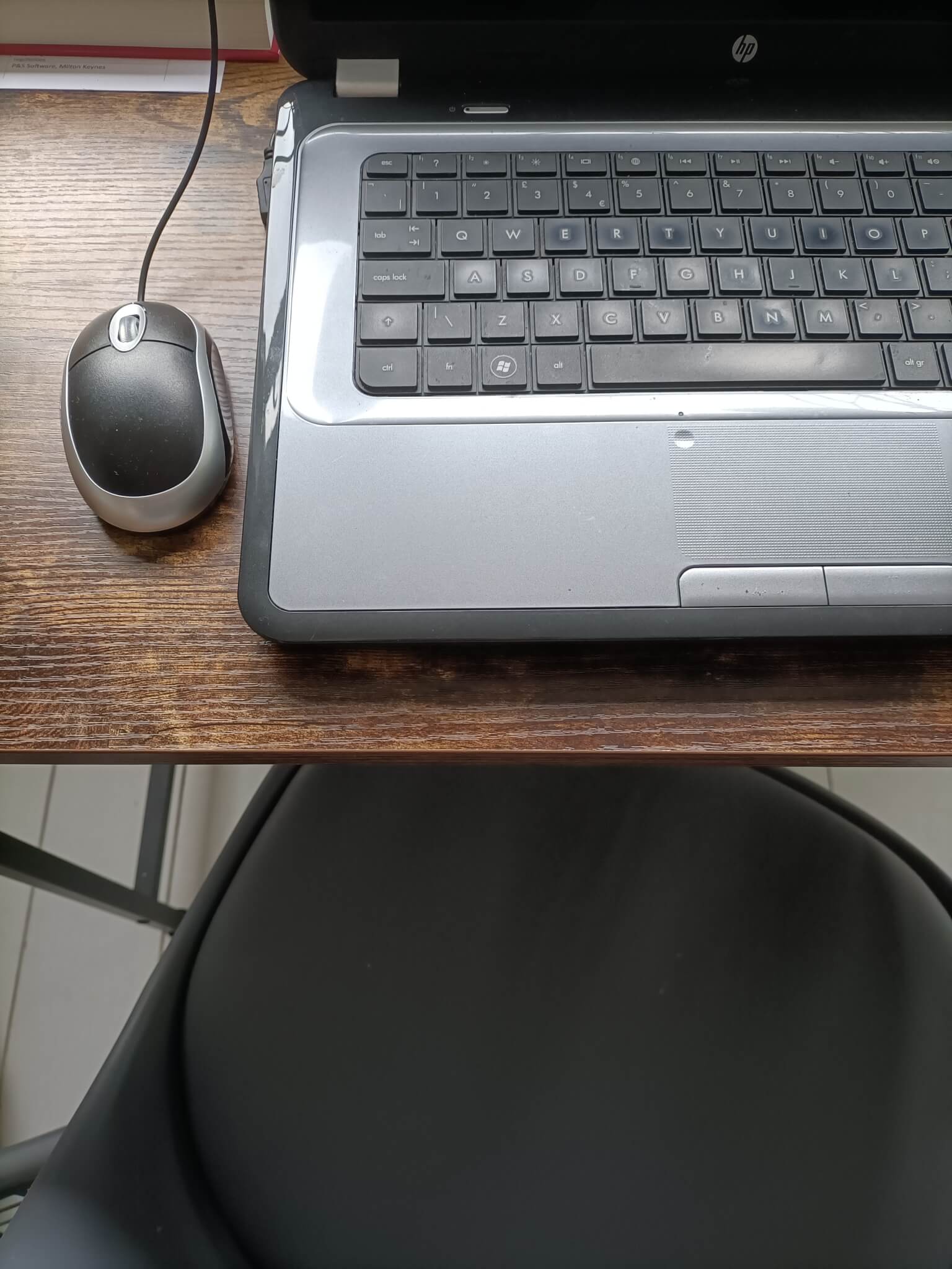 A laptop and wired mouse on a wooden desk, with a black chair in front. - Home Instead