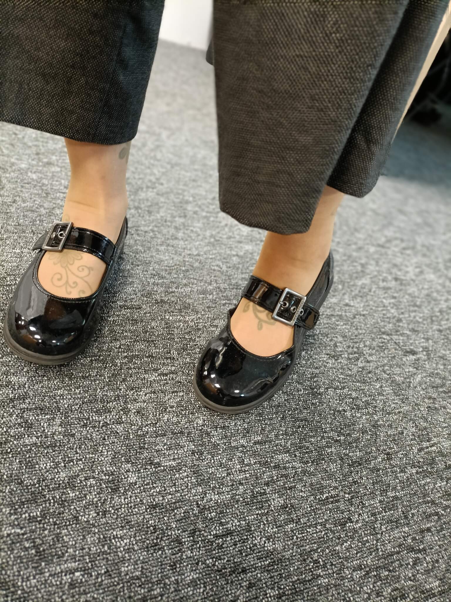 Close-up of a person wearing black Mary Jane shoes with ankle tattoos, standing on a gray carpeted floor. - Home Instead