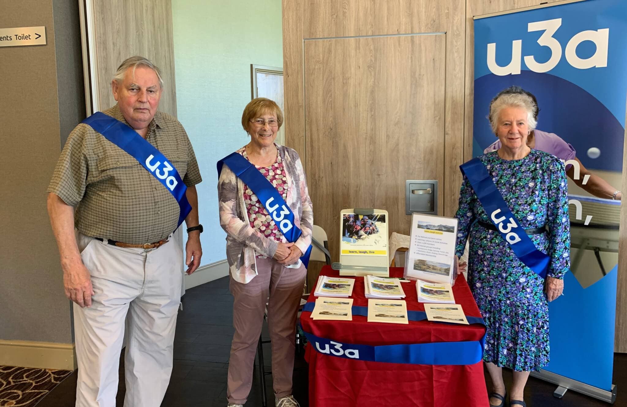 Three people wearing "u3a" sashes stand beside a table displaying brochures and flyers at an event. - Home Instead