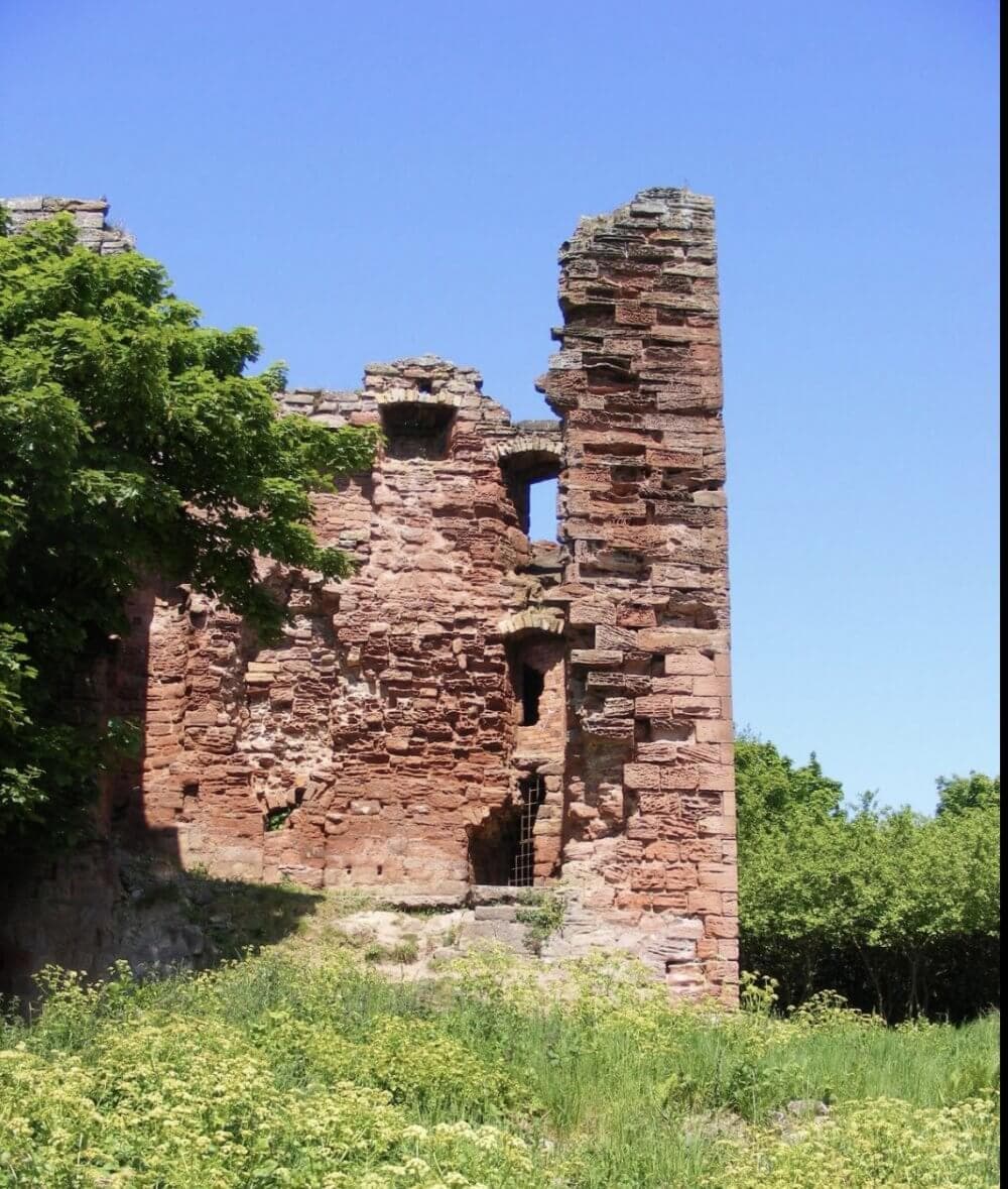 A crumbling red brick ruin stands against a clear blue sky, surrounded by green foliage and grass. - Home Instead