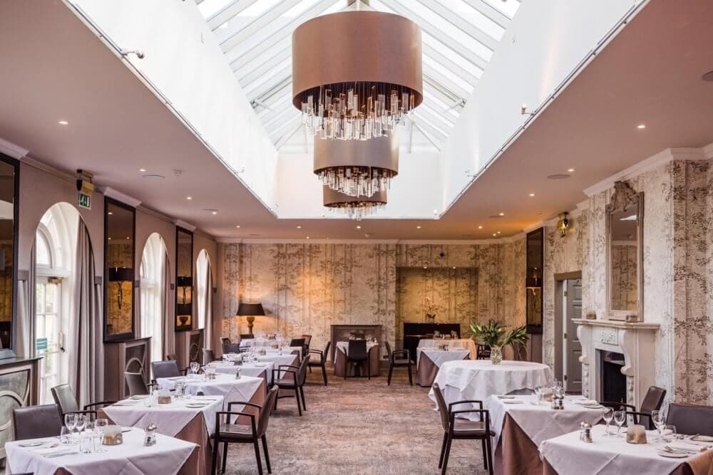 Elegant restaurant interior with skylight, chandeliers, arched windows, and tables set with white tablecloths and chairs. - Home Instead