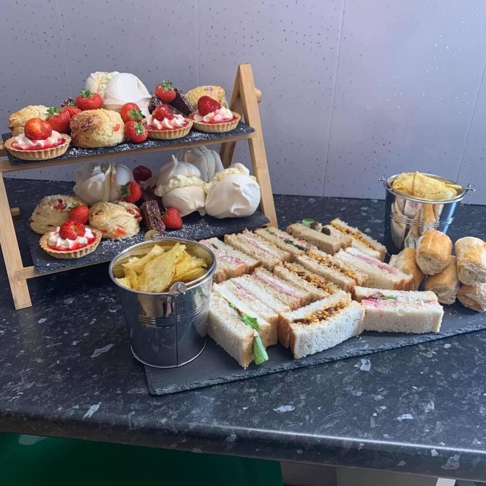 A tiered stand with scones and desserts, and a tray of sandwiches, crisps, and sausage rolls on a black countertop. - Home Instead