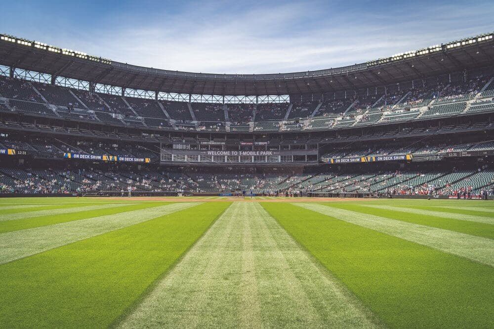 Wide-angle view of a baseball stadium from the outfield, with green grass and empty seats filling the stands. - Home Instead