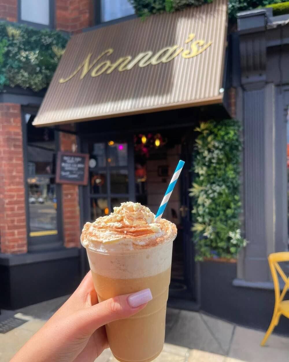 A hand holding a whipped cream-topped iced coffee in front of a café with a sign that reads "Nonna's". - Home Instead