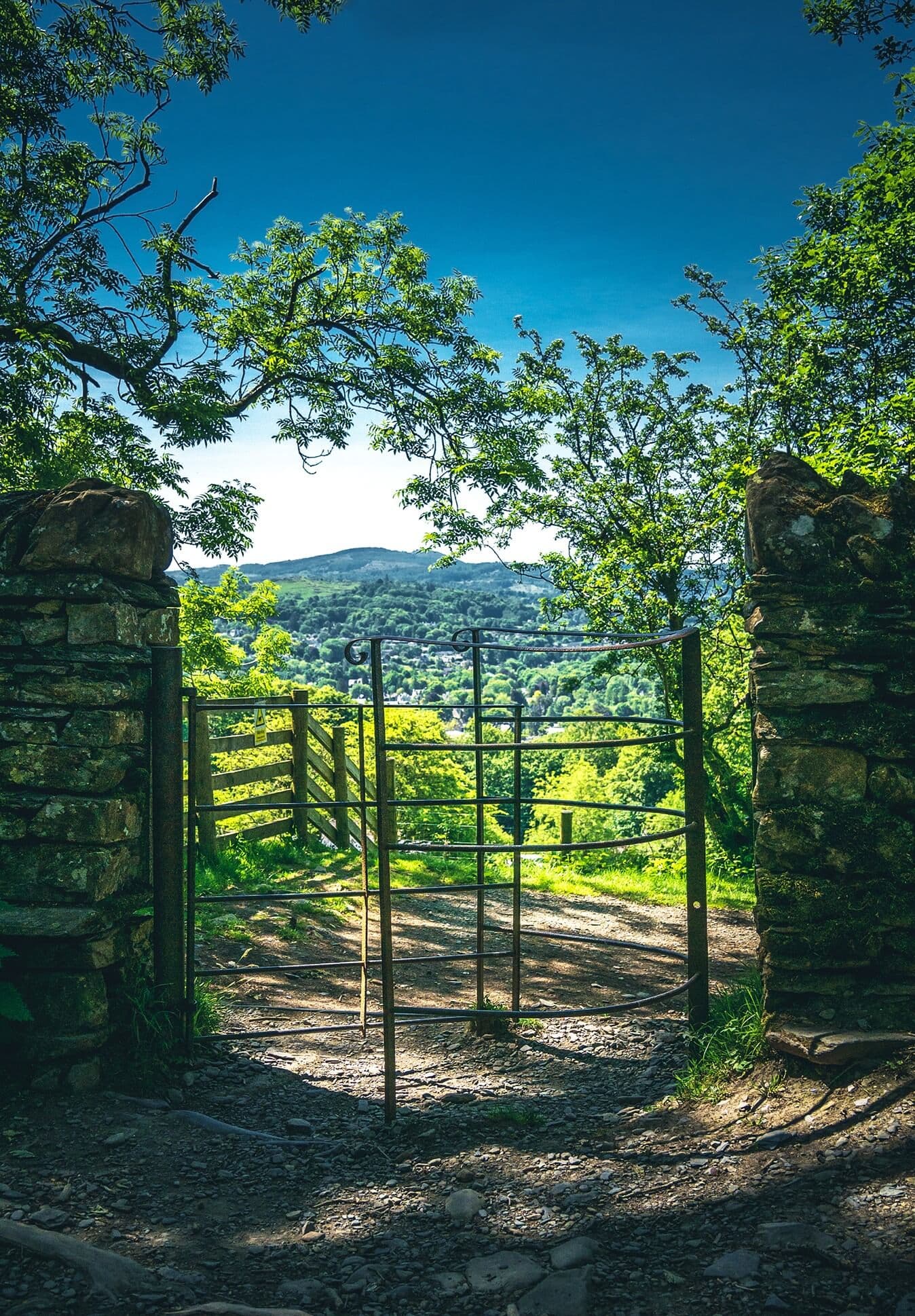 A scenic countryside view with a metal gate between stone pillars, trees, and hills in the background on a sunny day. - Home Instead