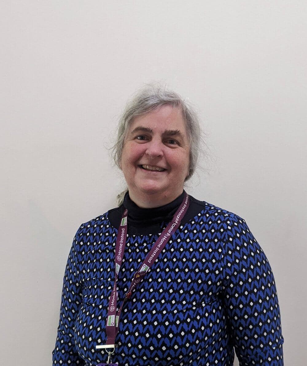 Smiling person with gray hair wearing a blue patterned shirt and a maroon lanyard against a neutral background. - Home Instead