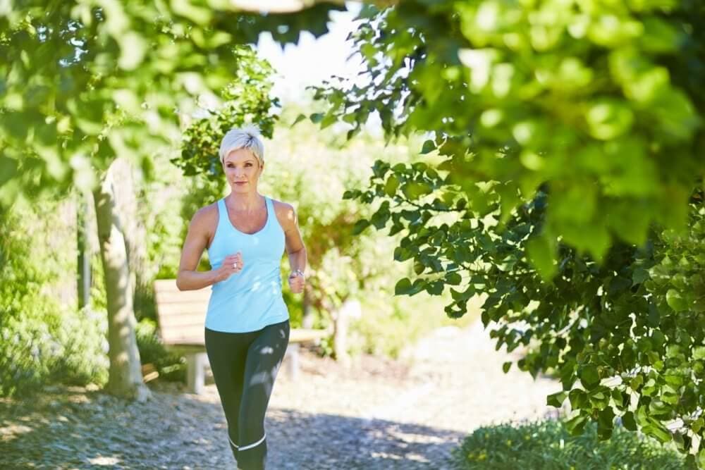 A woman in a blue tank top jogging on a leafy path on a sunny day, with vibrant green foliage around her. - Home Instead Bournemouth & Christchurch