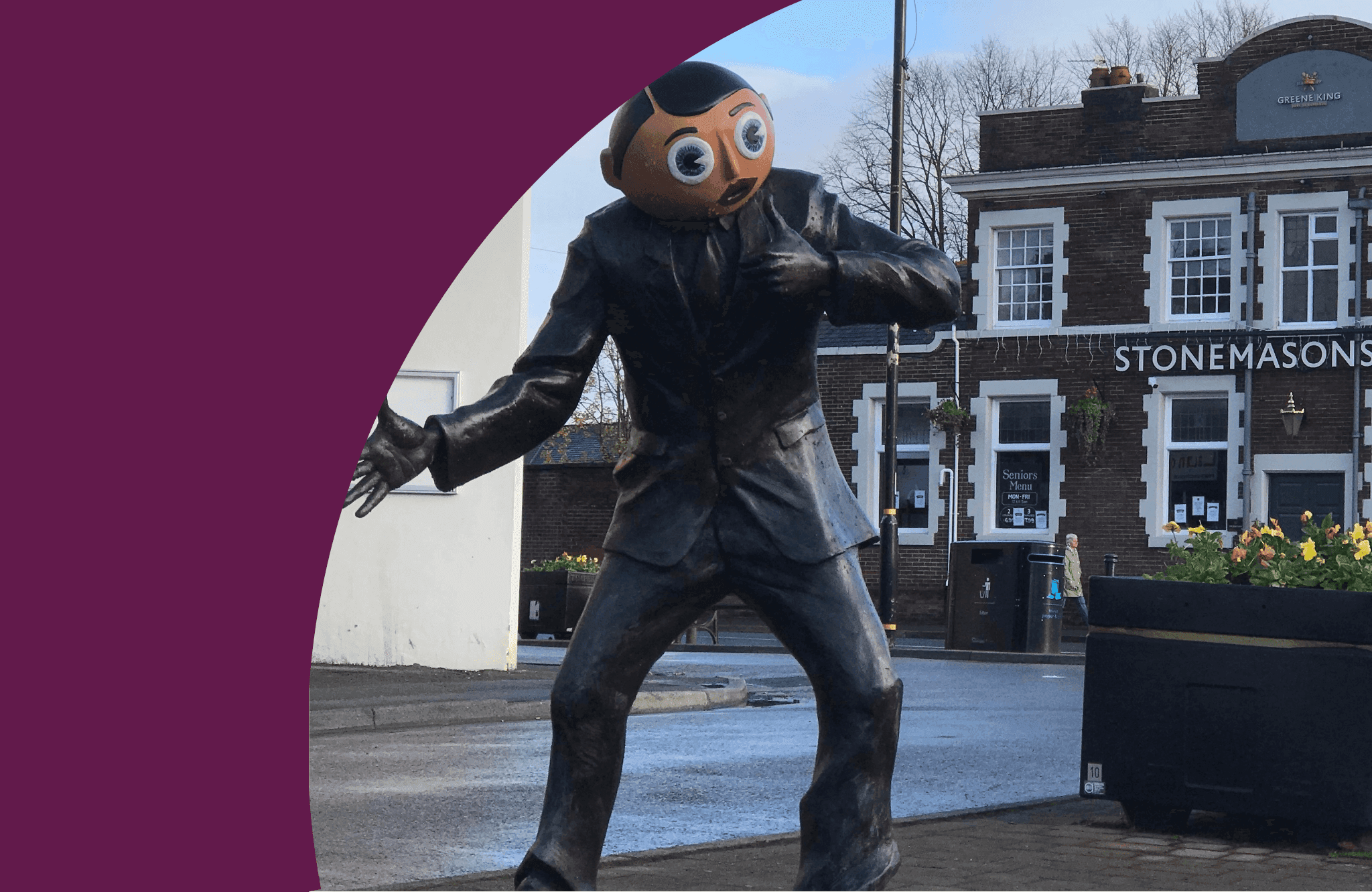 Statue of a person with a large, round head in a suit, positioned outdoors near buildings and a street. - Home Instead